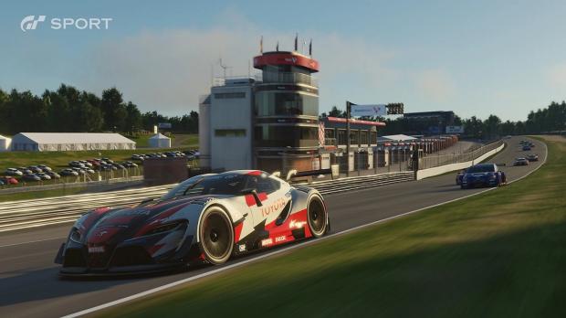 A Toyota race car rounds a track in Gran Turismo Sport, with other cars behind it and a pit building alongside the circuit.